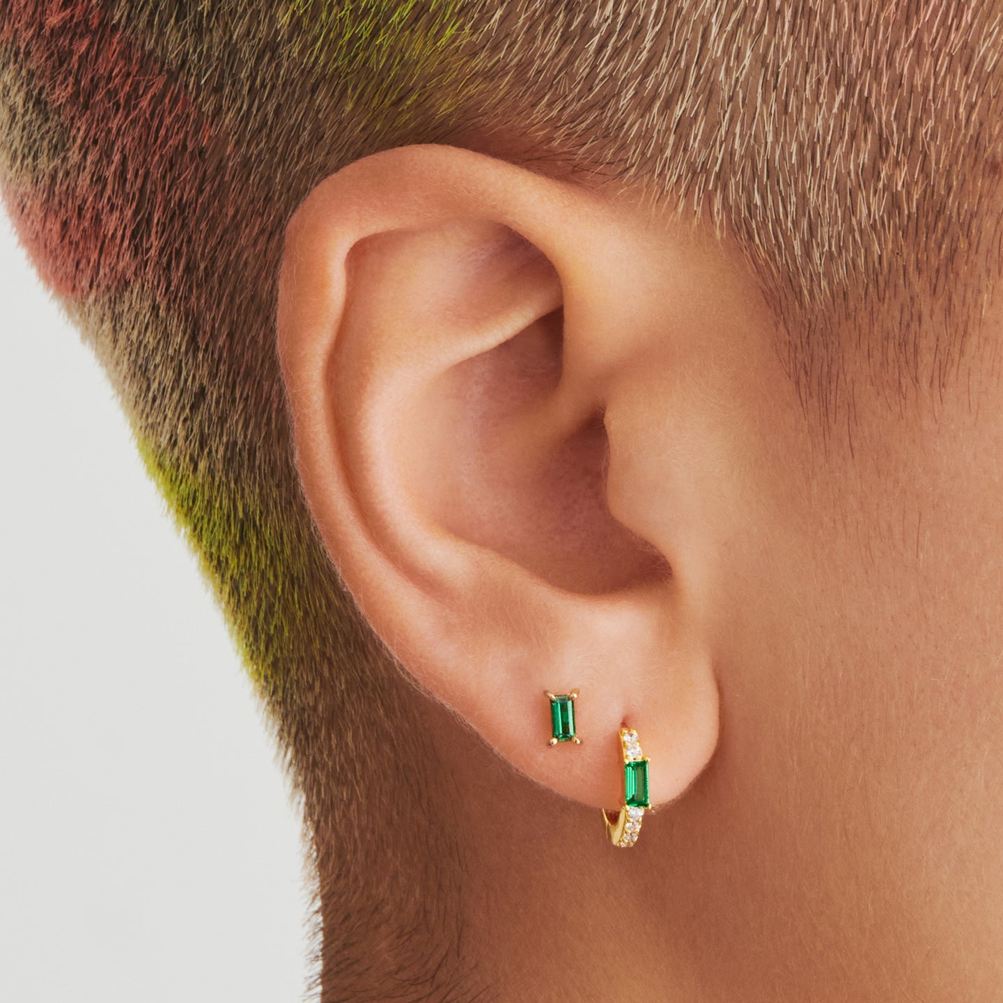 An image of a gold/clear huggie with a green accent baguette CZ stone on ear. [hover] color:null|gold/green