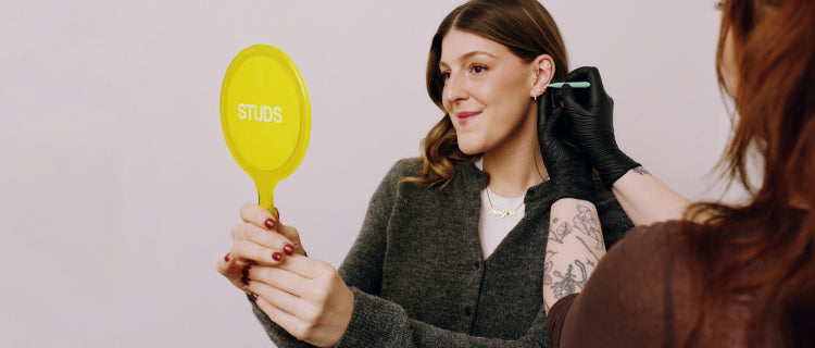 A woman getting her ear pierced while holding a yellow mirror.
