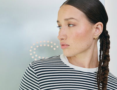 Woman with braided hair wearing a striped shirt against a light background with multiple ear piercings.