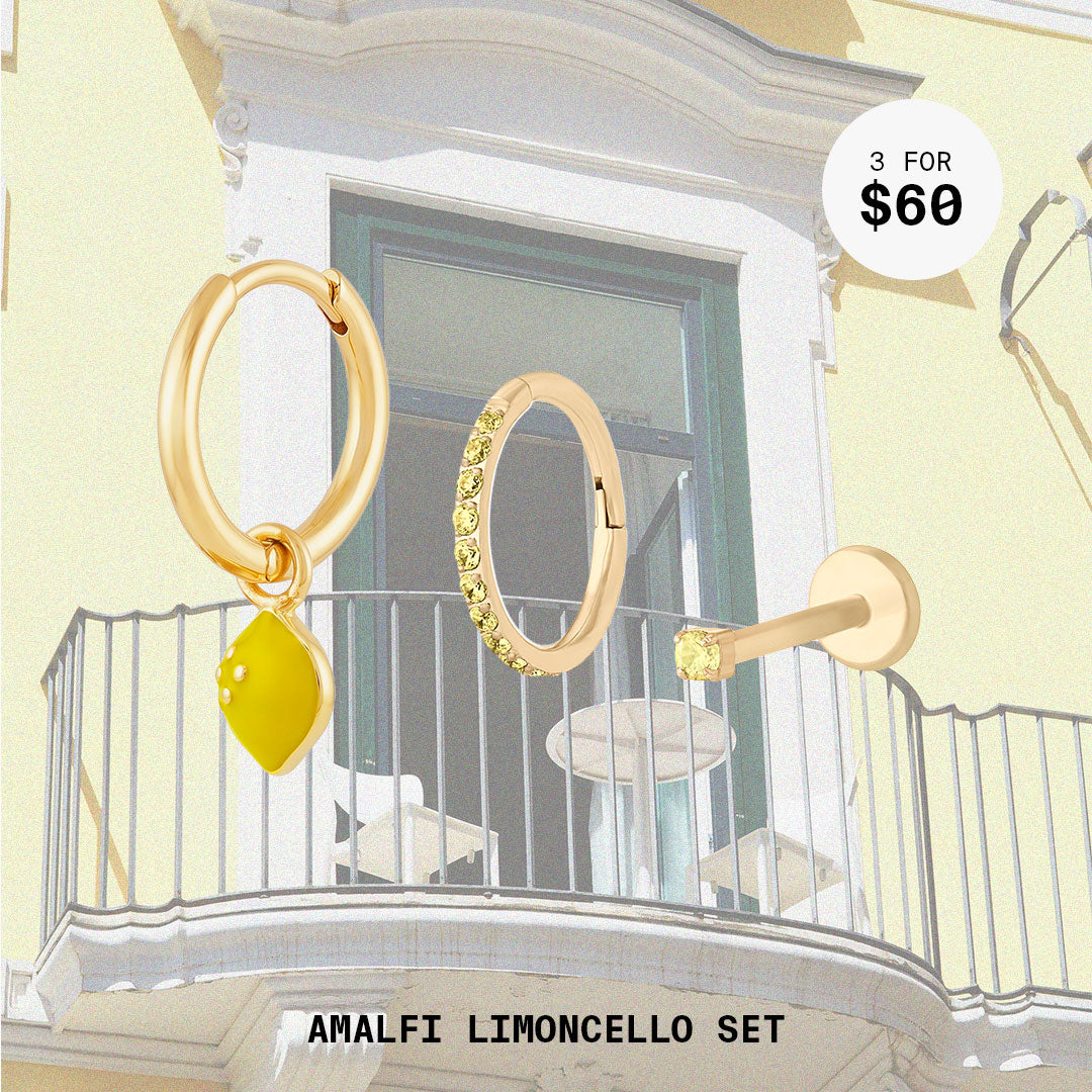 This image shows a balcony background with a set of three earrings. This set contains a lemon charm huggie, topaz pave clicker, and micro topaz flatback stud. All of these earrings are 14k gold plated.