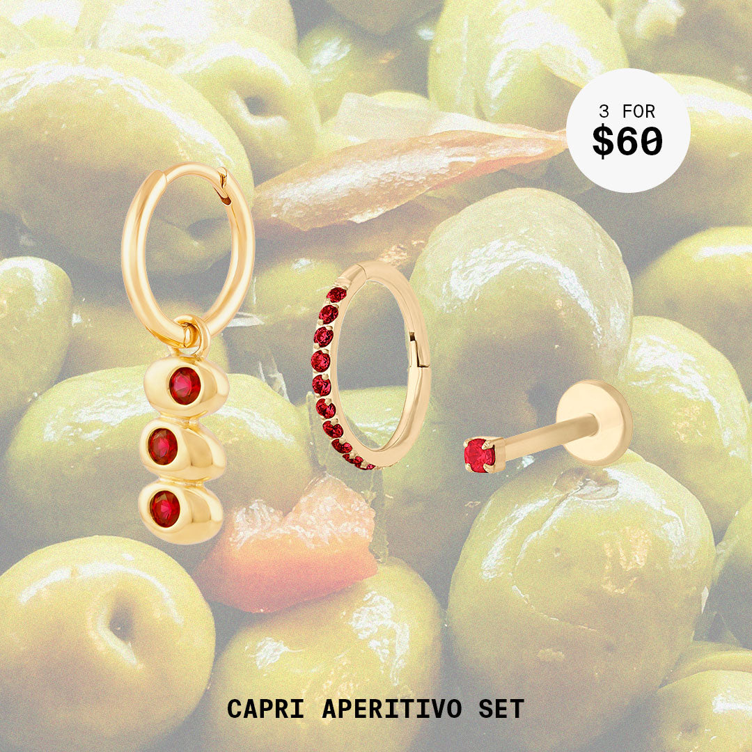 This image shows a background with olives and three earrings. The set includes an olive charm huggie, garnet pave clicker, and micro arnet flatback stud. These earrings are all 14k gold plated.