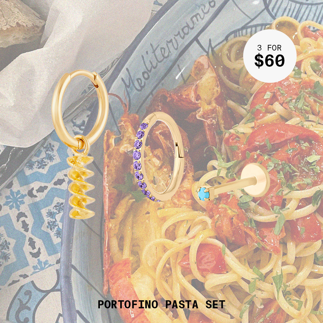 This image shows a background with a bowl of pasta and a set of three earrings. The set includes a pasta charm huggie, amethyst pave clicker, and micro turquoise flatback stud. All earrings in this set are 14k gold plated.