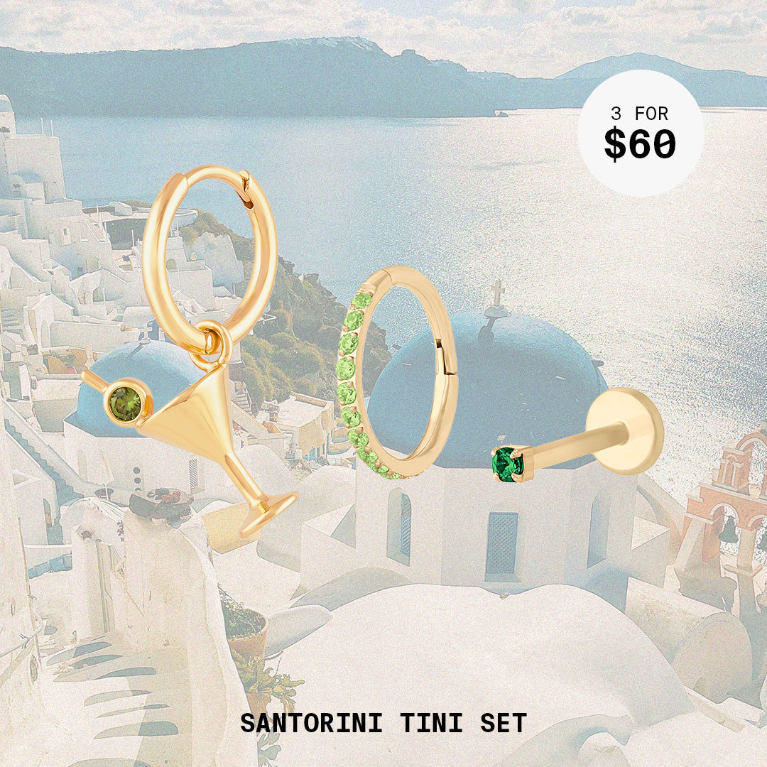This image shows a background of santorini and a set of three earrings. The set includes a martini charm huggie, peridot pave clicker, and emerald micro flatback stud. All earrings in this set are 14k gold plated.