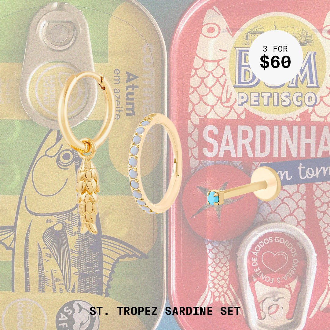 This image shows a background with sardine cans and a set of three earrings. The set includes a fish charm huggie, pearl pave clicker, and micro turquoise flatback stud. All earrings in this set are 14k gold plated.