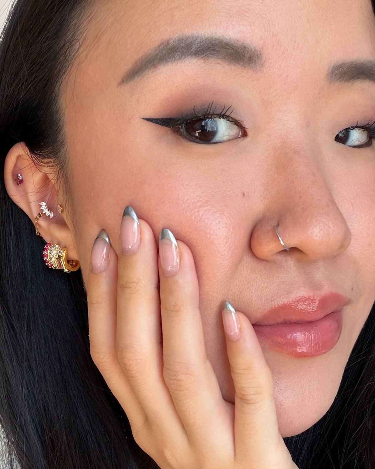 Close-up of a person's face and hand showcasing multiple earrings, including a silver hoop in a nose ring, a cluster of silver and pink earrings on the ear lobe, and silver-tipped French-tail manicured nails.