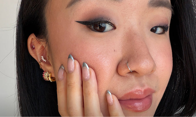 Close-up of a person wearing multiple earrings, including a gold hoop with a pink gem, a gold ring with a pearl-like center, and a silver huggie hoop, along with a silver nose ring.
