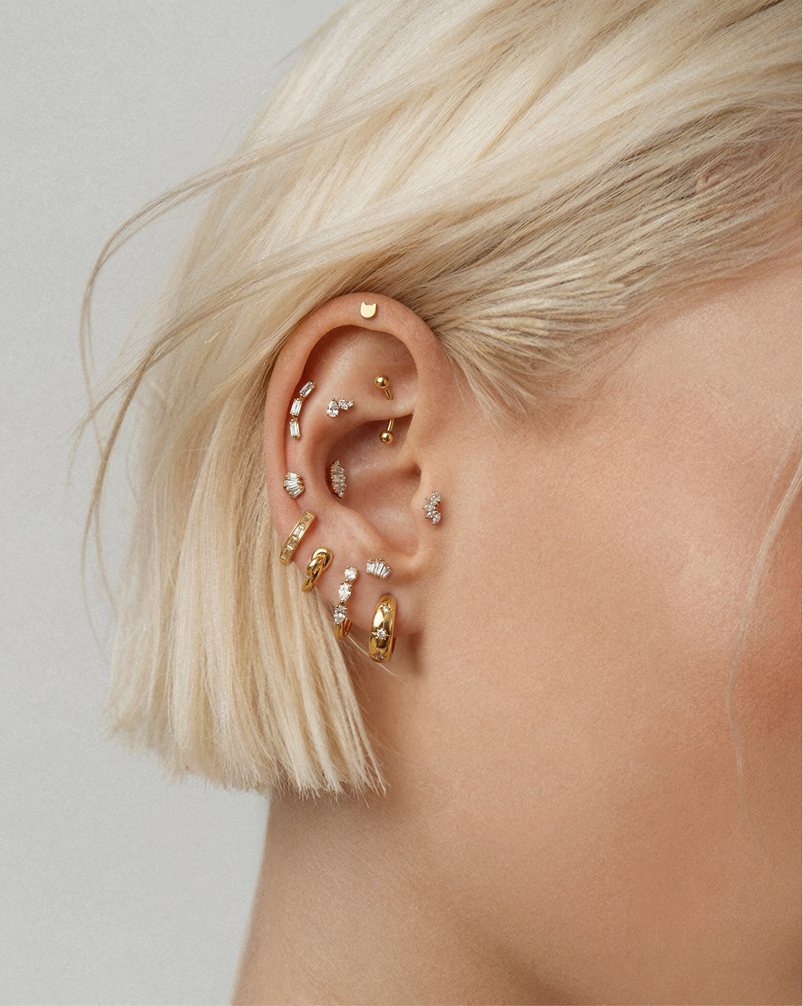 Close-up of a person's ear adorned with multiple gold and silver ear piercings and small clear stones, with short platinum blonde hair against a light gray background.