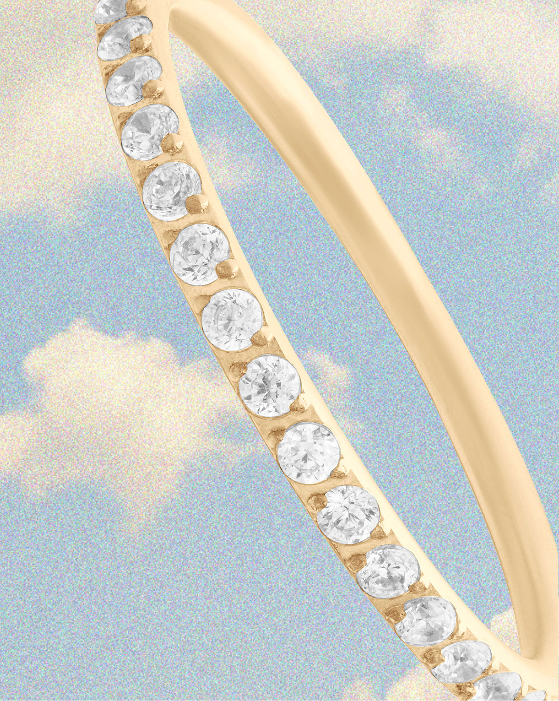 A close-up of a yellow gold ring featuring a row of round clear gemstones along the band, set against a light blue sky background with soft clouds.