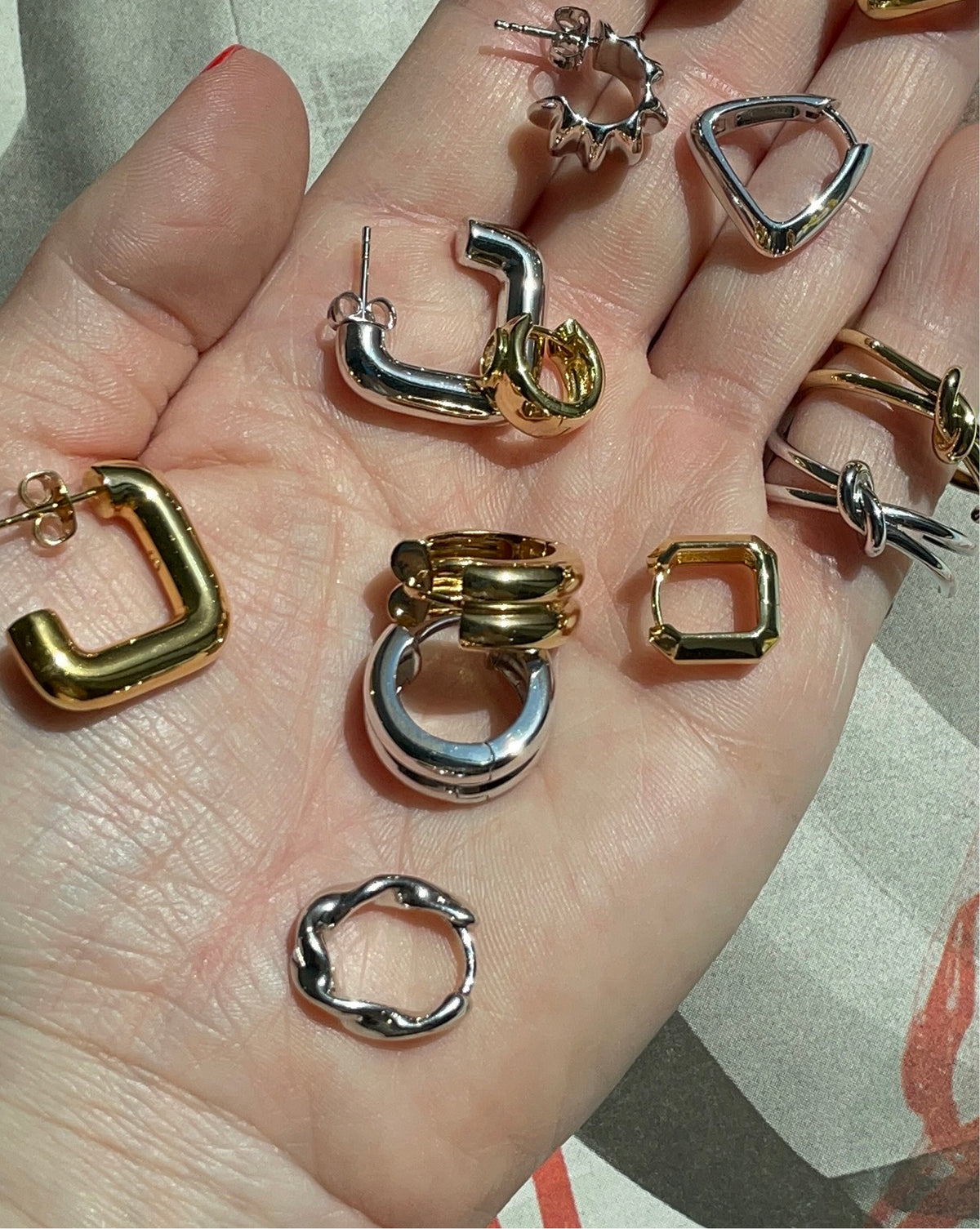 A hand holding multiple abstract geometric earrings in silver and gold finishes, including hoop, square, and twisted designs.