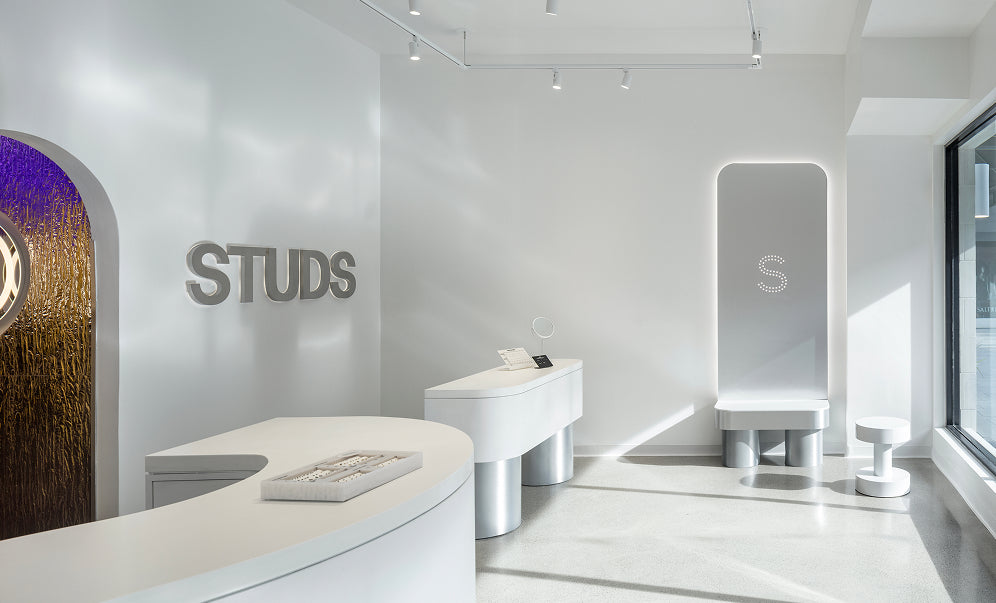 A modern, minimalist beauty studio interior with white furniture, a curved reception desk, a large arched mirror, and the word 'STUDS' mounted on the wall.