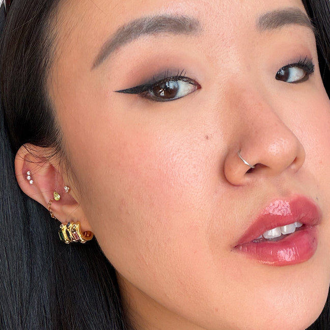 Close-up of a person's ear showcasing multiple gold and silver cartilage piercings, including a small hoop, a cluster of studs, and a larger stacked barelore. The person has dark hair, winged black eyeliner, and glossy pink lips.