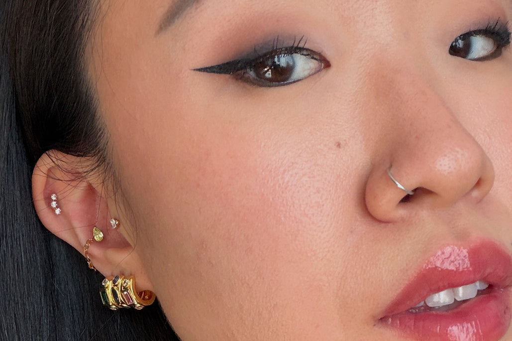 Close-up of a person's ear showcasing multiple gold and silver piercings, including a prominent stacked gold hoop and several smaller studs, with a nose ring on the nose.