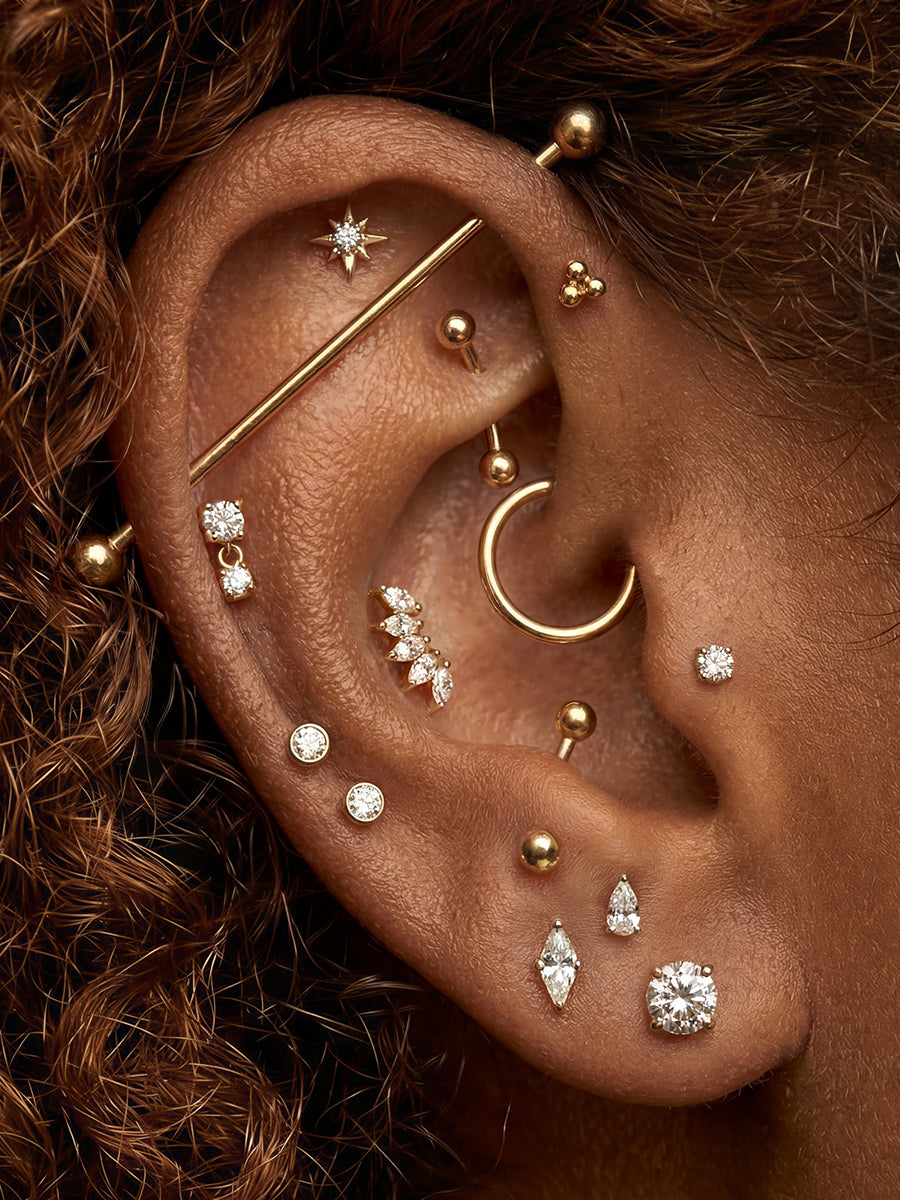 Close-up of a human ear wearing multiple gold ear piercings and earrings, including a hoop, barbell, studs, and crystal-like accents, against dark brown hair.