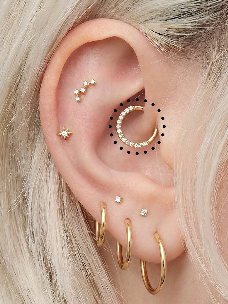 Close-up of a person's ear showcasing multiple gold ear piercings, including a large crescent hoop with clear rhinestones, several small studs, and hoop earrings, with blonde hair framing the ear.