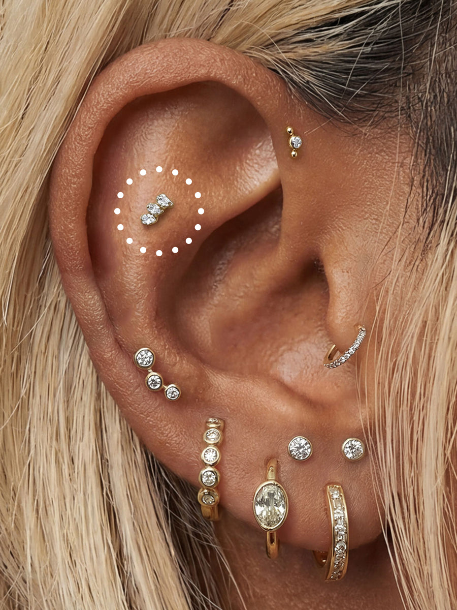 Close-up of a person's ear adorned with multiple gold and silver ear piercings and earrings, including a small silver stud in the helix, a curved gold hoop in the lobe, and several gold and silver cartilage piercings. Blonde hair frames the ear, and a white dotted circle highlights one of the ear piercings.
