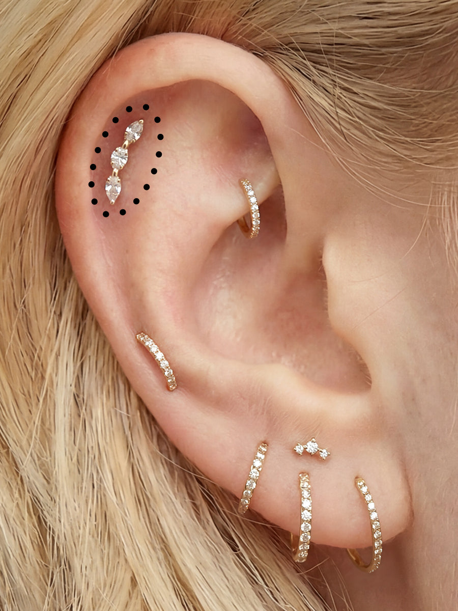 Close-up of a person's ear wearing multiple delicate gold ear cuffs and studs, with black dotted placement markers indicating where additional earrings could be worn.