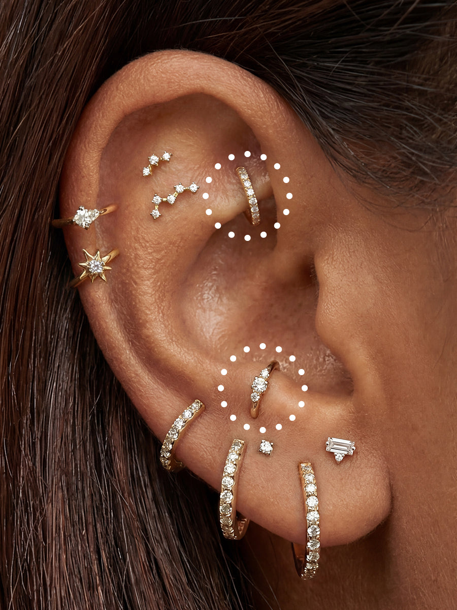 A close-up of a person's ear adorned with multiple gold and silver cartilage piercings and studs, including several small clear gemstone accents. White dotted circles highlight different jewelry placements on the ear.