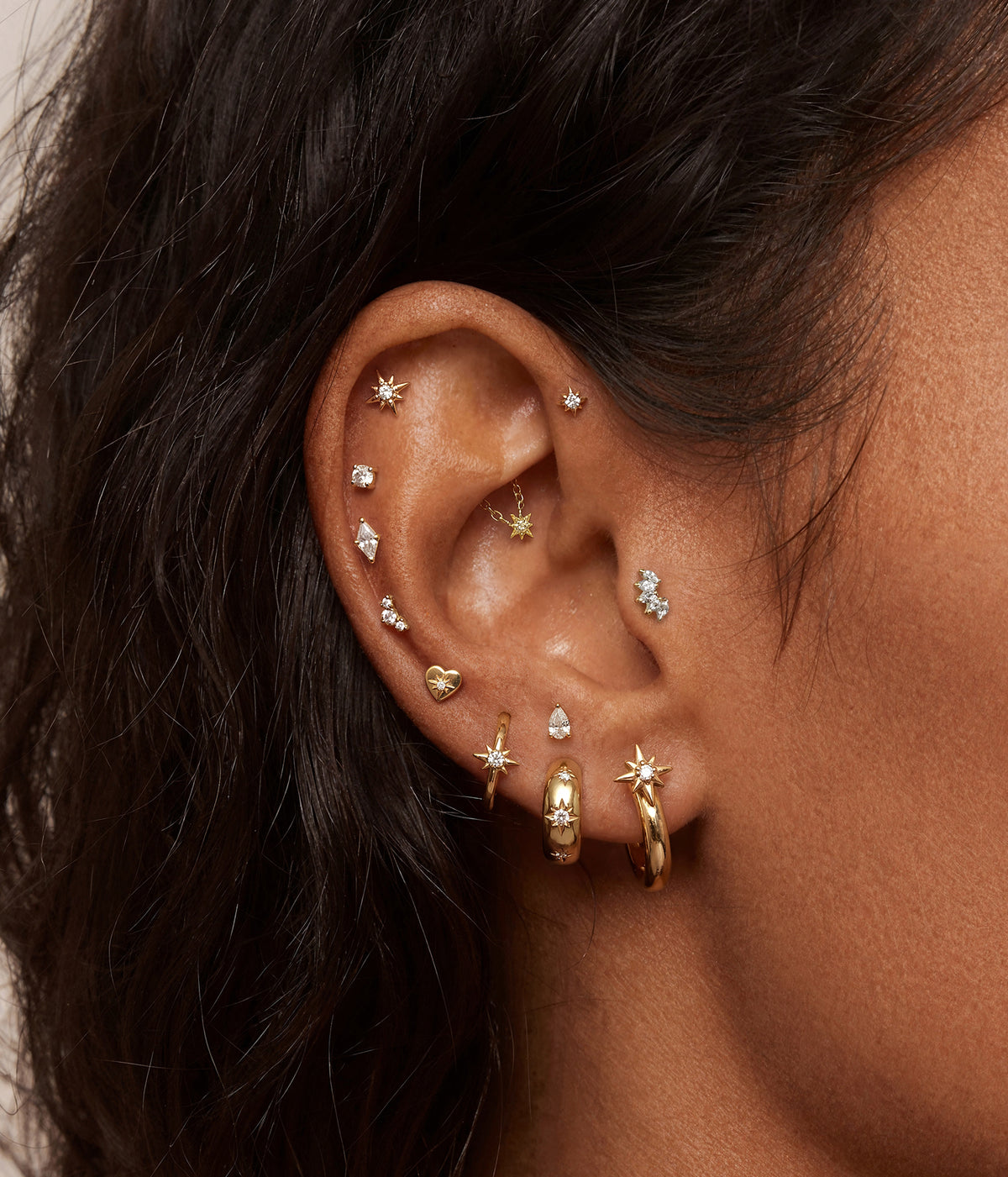 A close-up of a person's ear adorned with multiple gold and silver ear piercings, including star-shaped studs, hoop earrings, and small geometric studs.