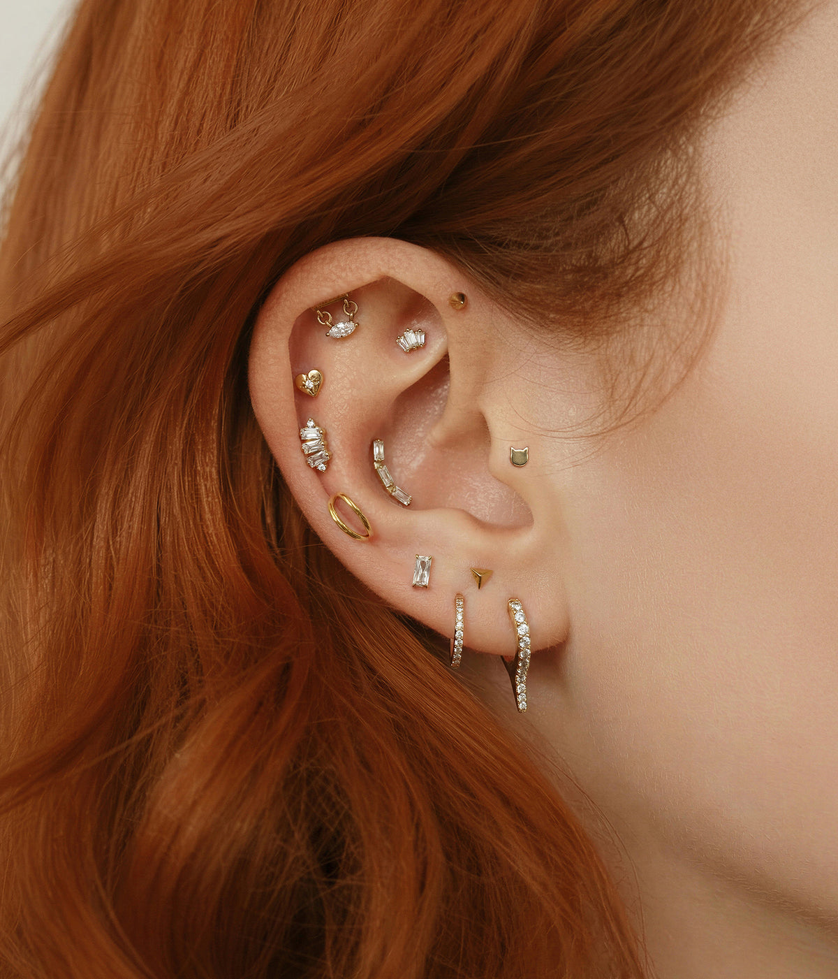 A close-up of a person's ear adorned with multiple gold and silver ear piercings and earrings, with red hair framing the side of the face.