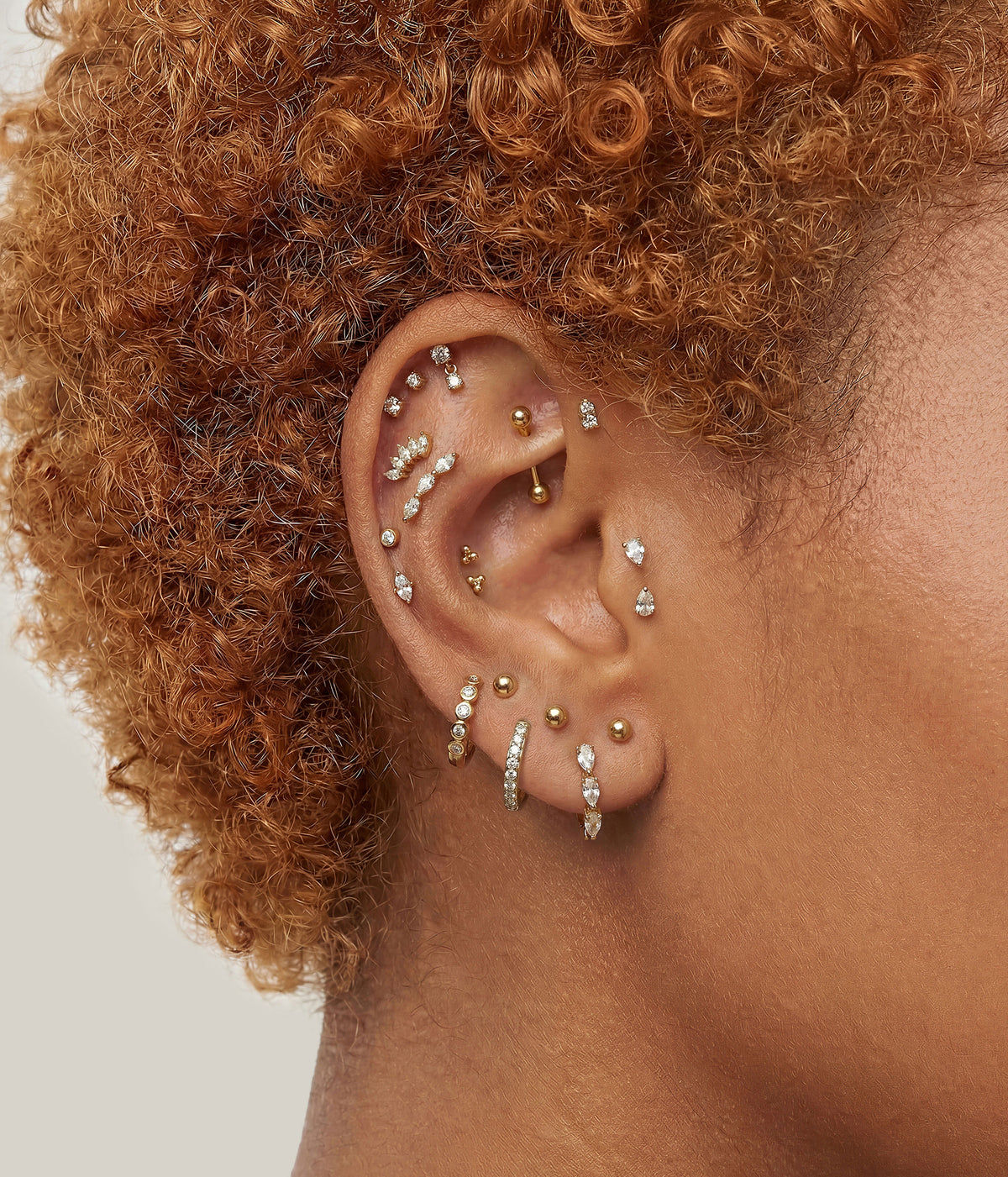 A close-up of a person's ear adorned with multiple gold and silver ear piercings and stud earrings, featuring curly auburn hair against a neutral light beige background.