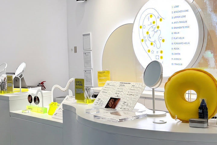 A modern jewelry display counter with a large white circular mirror, a yellow donut-shaped piercing pillow, small mirrors, and earrings displayed on a white curved counter in a bright interior.