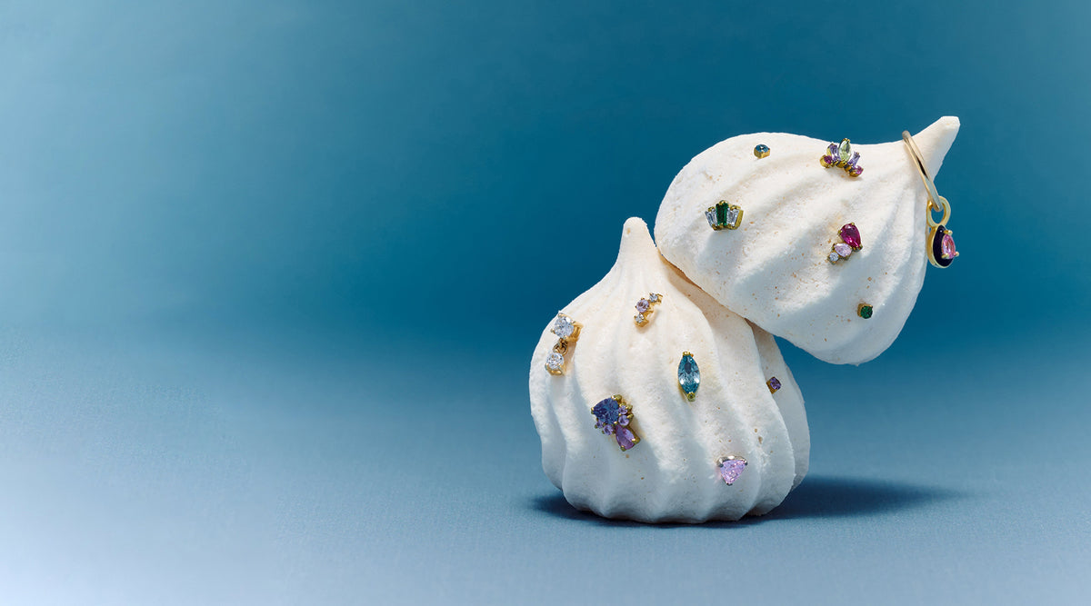 Two white meringue candies decorated with multiple colorful gemstones earrings from the new candy collection photographed against a smooth blue gradient background.