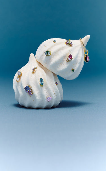 Two white meringue candies decorated with multiple colorful gemstones earrings from the new candy collection photographed against a smooth blue gradient background.