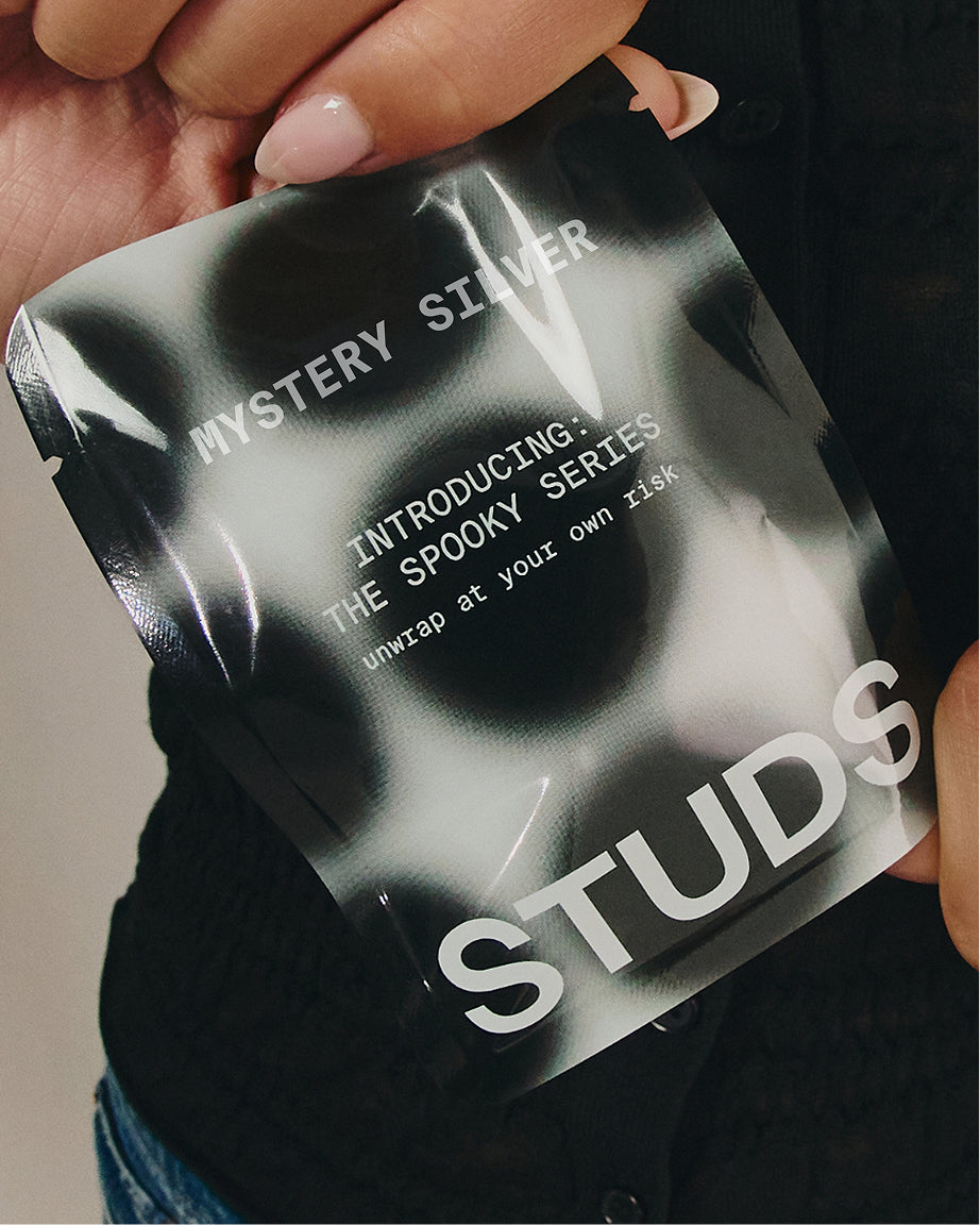 A close-up of a person holding a glossy black and white pouch labeled 'MYSTERY SILVER' and 'STUDS', with visible hands and dark clothing in the background. 