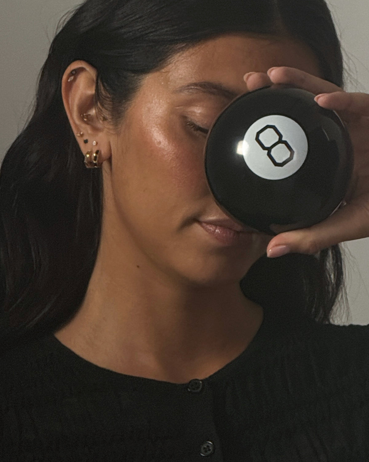 A close-up lifestyle shot of a woman holding a black 8-ball in front of her eye, wearing a black knit top and small gold hoop earrings against a neutral gray background.