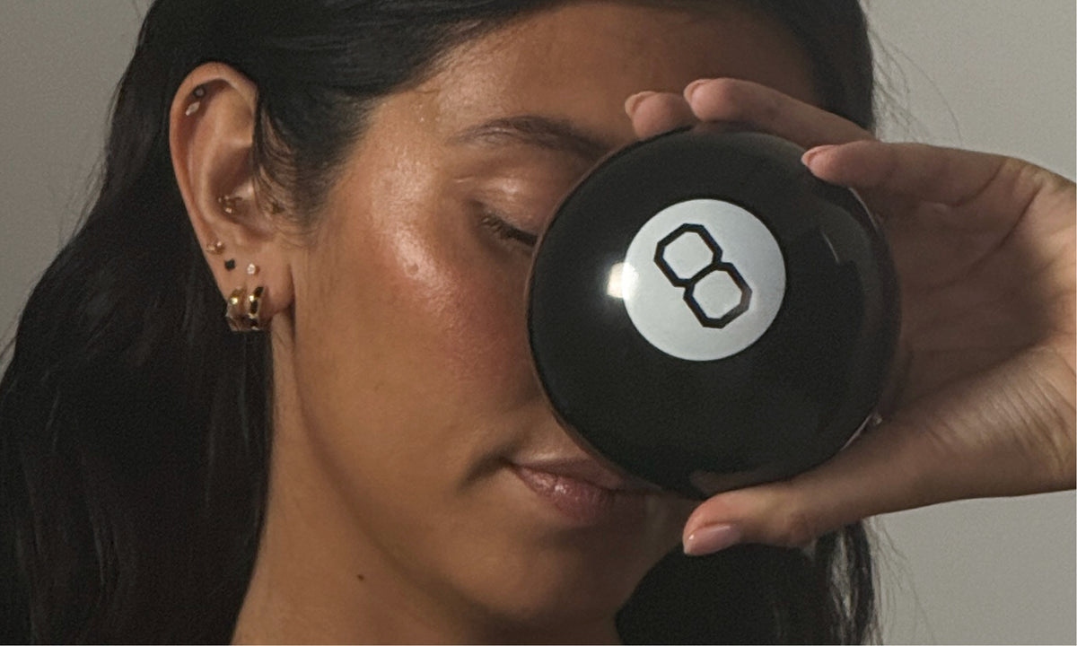 A close-up lifestyle shot of a woman holding a black 8-ball in front of her eye, wearing a black knit top and small gold hoop earrings against a neutral gray background.