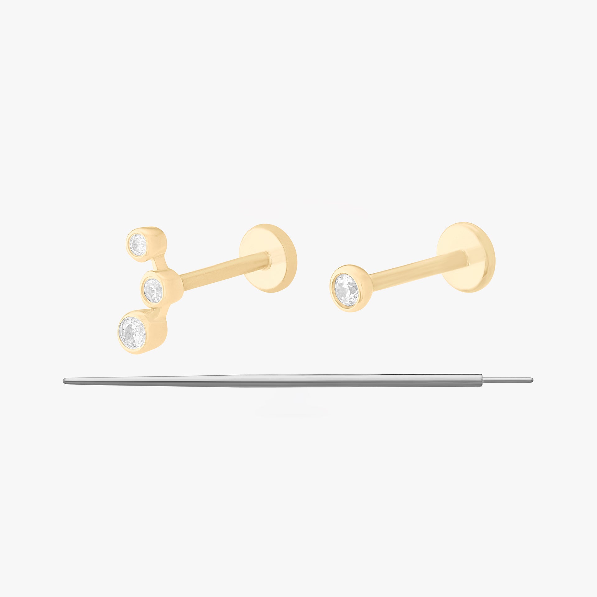 This image displays a set of two 14K gold flatback studs and a flatback insertion tool. The two earrings included in this set are the 14K Bezel Flatback Stud in gold/clear, as well as the 14K CZ Bezel Cluster Flatback Stud in gold/clear.