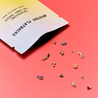 A  gradient bag labeled 'MYSTERY FLATBACKS' open on a coral pink background, with several small flatback earrings scattered out of it.