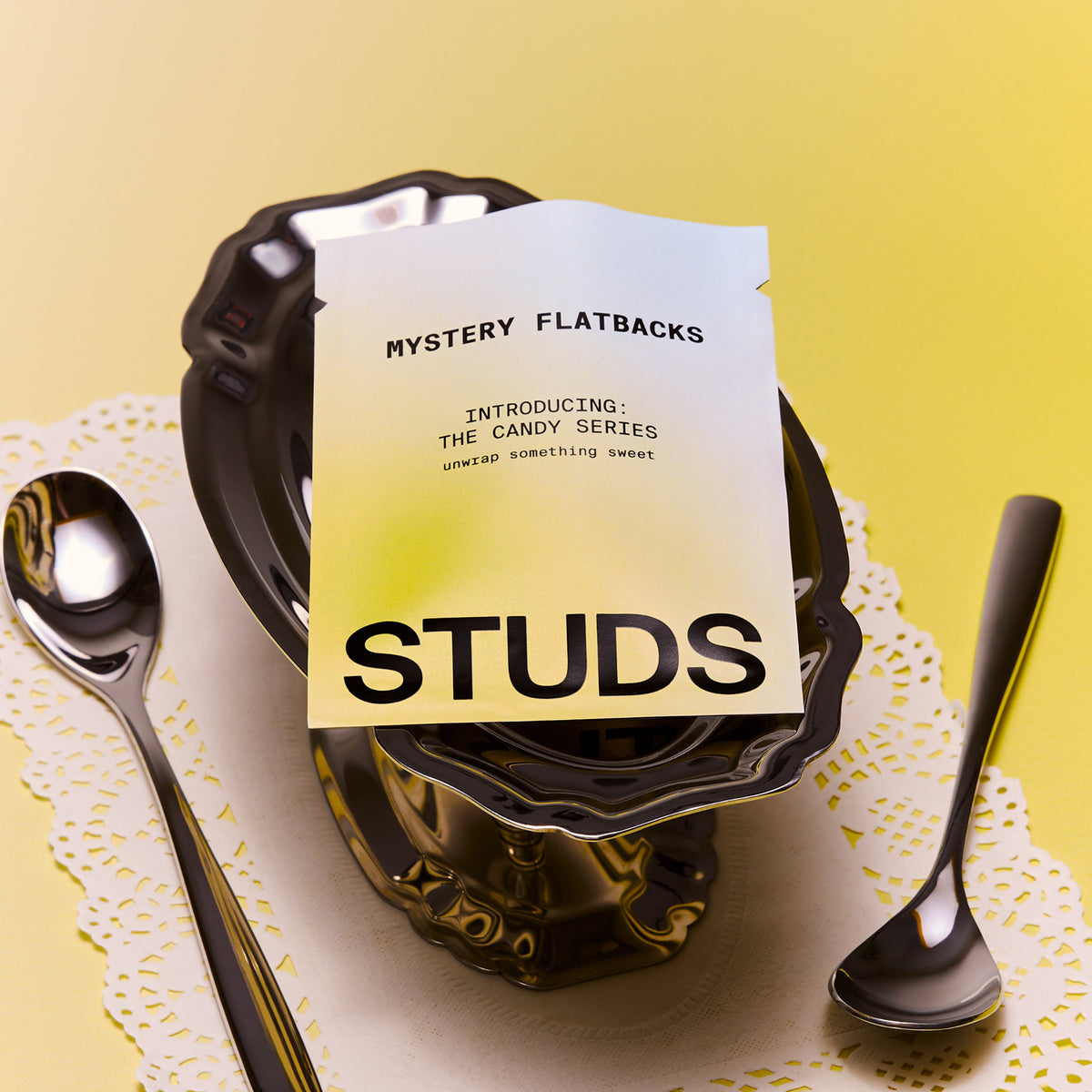A flatback candy packet labeled 'MYSTERY FLATBACKS' and 'STUDS' is placed on a bowl with two spoons on a yellow background.