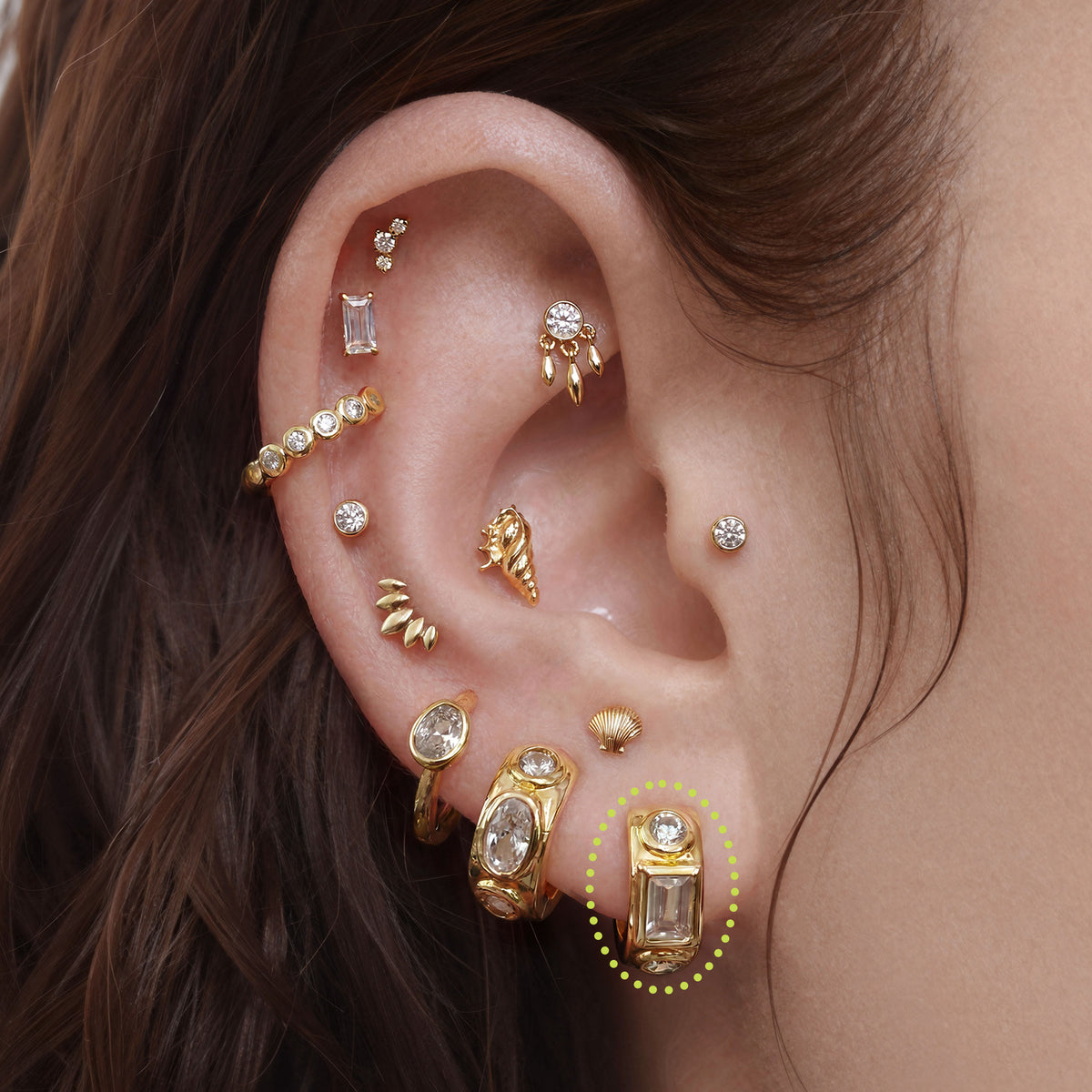 This image shows an ear with a 14k gold plated huggie with three clear cz stones. One square in the middle and two circular stones on the outside.