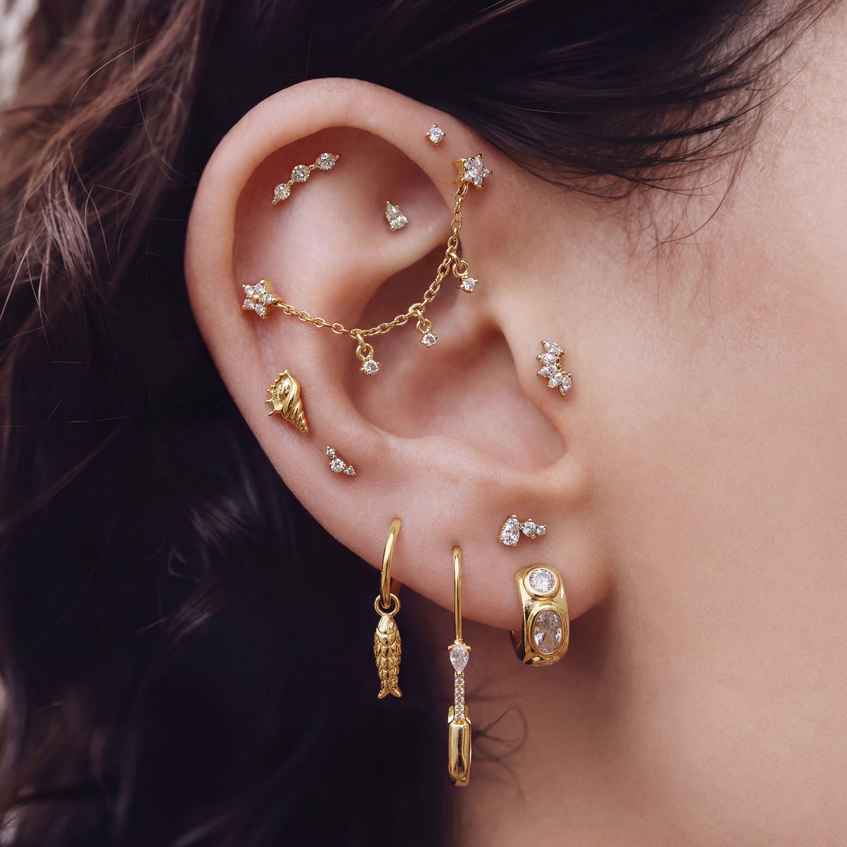 This is an image of an ear with a anodized gold titanium chained flatback stud with two flatback posts. Each flatback stud is a clear star CZ, and there is a chain with three clear CZ stones that connect the two earrings. There is also a huggie with a fish charm on the lobe. 