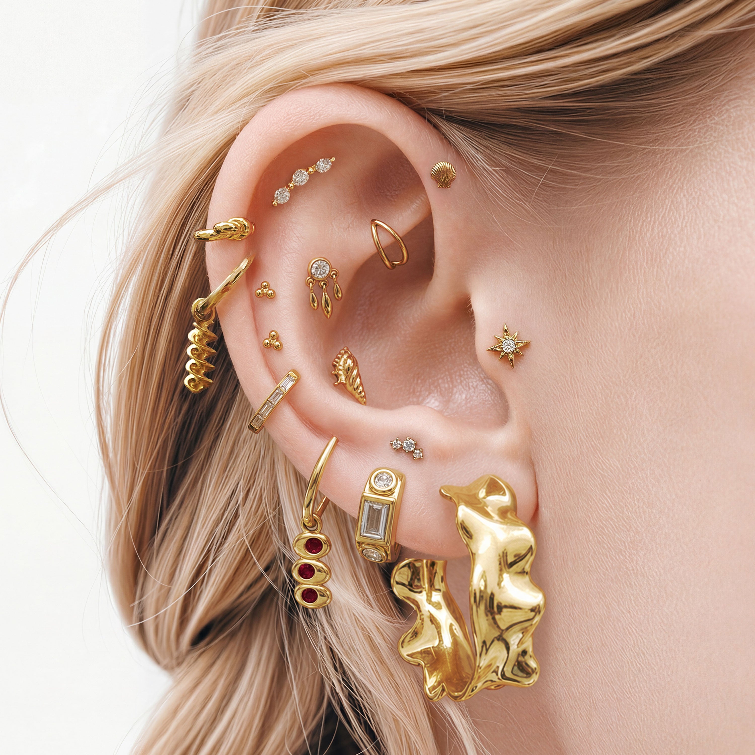 This image shows an ear with a 14k gold plated huggie with a spiral pasta charm  and a huggie with an olive charm with red gemstones. 