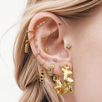 This image shows an ear with a 14k gold plated huggie with a spiral pasta charm  and a huggie with an olive charm with red gemstones. 