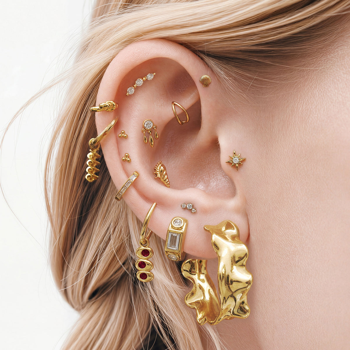 This image shows an ear with a 14k gold plated huggie with a spiral pasta charm  and a huggie with an olive charm with red gemstones. 