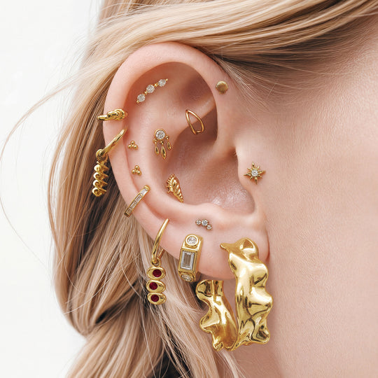 This image shows an ear with a 14k gold plated huggie with a spiral pasta charm  and a huggie with an olive charm with red gemstones. 