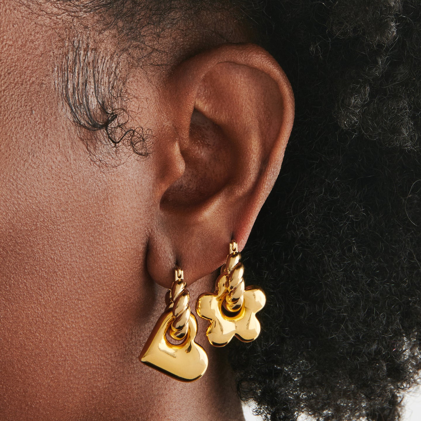 a gold earring set that includes two small french twist hoops, a puffy daisy charm, and a puffy heart charm on ear [hover] color:null|gold