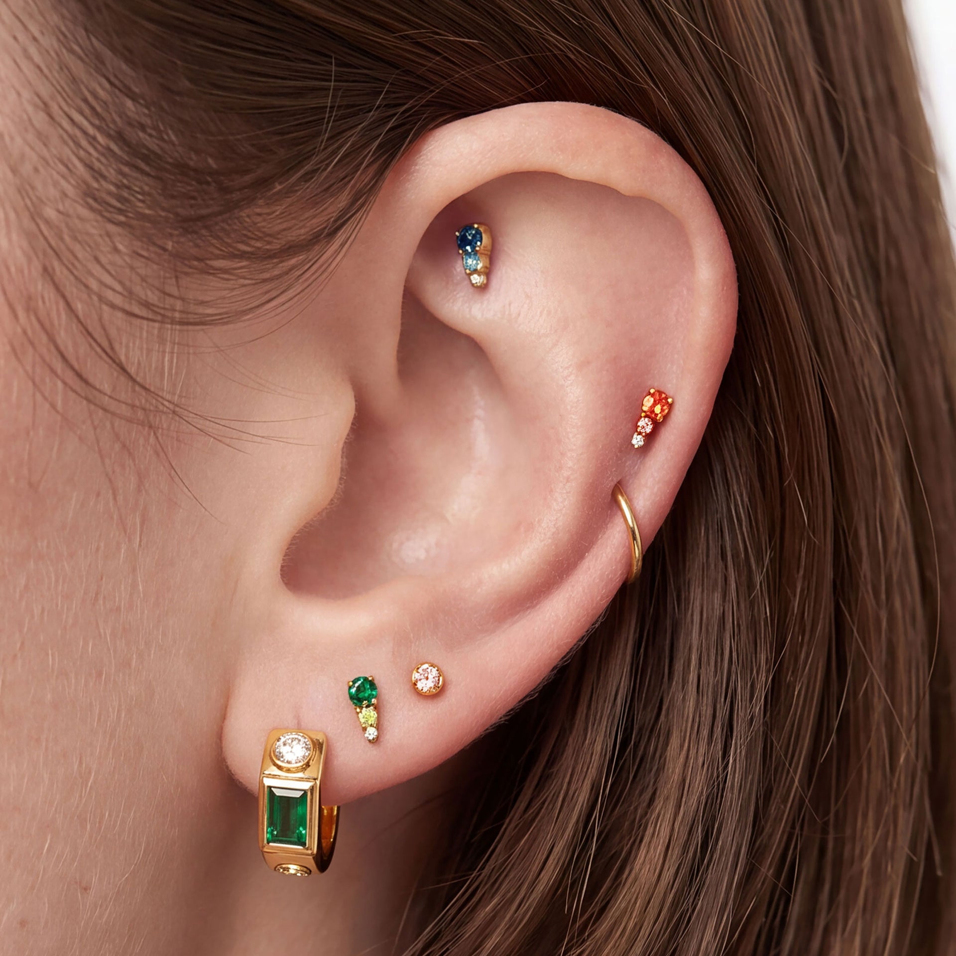 This image shows an ear with the three  golden titanium flatback studs with color cz stones. There is a blue cz stone flatback, a pink cz stone flatback and a green cz stone flatback. - Alternative view 1