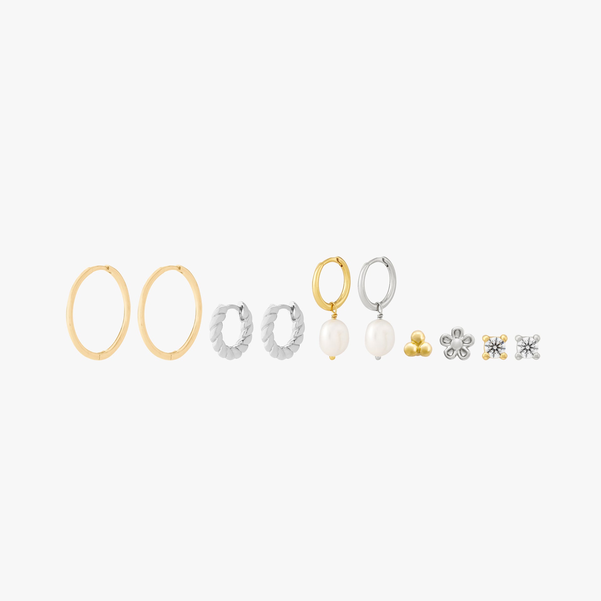 This image shows a set of 14k Yellow Gold and silver earrings. The set includes a gold pair of thing hoops, a silver french twist pair of huggies, one silver pearl huggie., one gold pearl huggie, one gold trinity ball stud flatback, a silver daisy flatback, one gold clear cz stone stud and one silver clear cz stone stud.