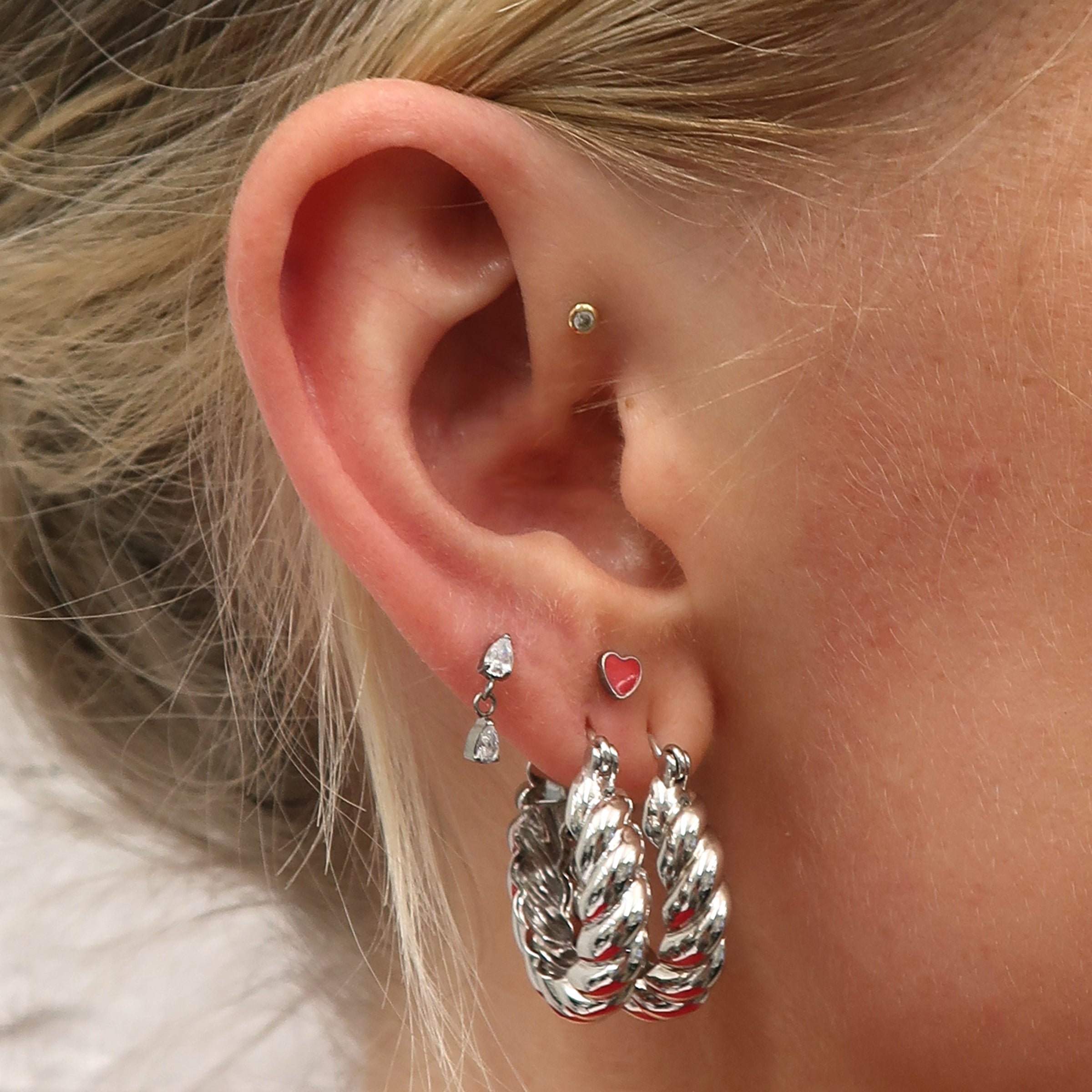 Close-up of an ear with two chunky silver hoops, a silver heart-shaped flatback with red enamel, and silver dangle flatback with clear pear shaped stones